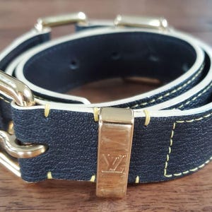 May include: A black leather belt with a gold buckle and white stitching. The belt has a gold LV logo on the buckle.