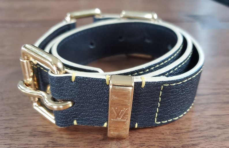 May include: A black leather belt with a gold buckle and white stitching. The belt has a gold LV logo on the buckle.