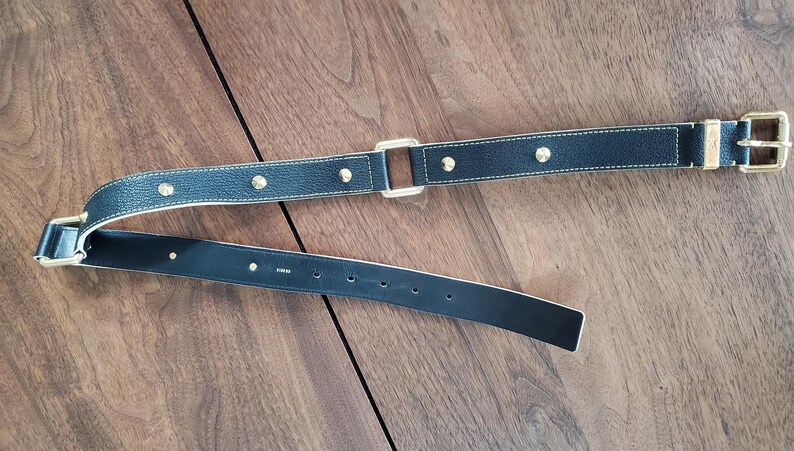 May include: A black leather belt with gold-toned hardware and a studded design. The belt is shown folded in half.