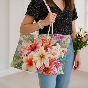 May include: A floral tote bag with a watercolour design of hibiscus and lily flowers in red, pink, and white. The bag has rope handles. The bag is held by a person wearing a black shirt and blue jeans.