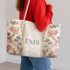 Op de afbeelding: Een grote tote bag met een crèmekleurige achtergrond en een bloemen- en Aziatisch geïnspireerd landschapsontwerp. De tas heeft de initialen "EMB" in teal en heeft crèmekleurige touwhandvatten.