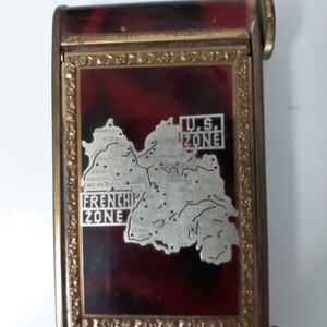 May include: A red and gold metal cigarette case with a map of Germany showing the U.S. Zone and the French Zone. The case has a hinged lid and a small latch.