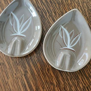 May include: Two teardrop-shaped ceramic spoon rests with a glossy taupe finish. Each piece features a white hand-painted botanical leaf design and a molded U-shaped indentation at the base to hold a spoon handle securely in place.