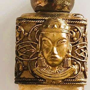 May include: A small, antique perfume bottle with a gold-colored metal casing. The bottle features an embossed face of a person with ornate detailing and a round stopper. The base of the bottle is clear.