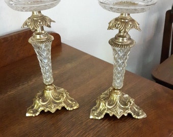 May include: A pair of gold metal candle holders with clear glass bases and decorative tops. The bases have a scalloped design and the tops have a floral design.