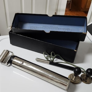 May include: An antique medical device with a silver metal body, black accents, and two black knobs. The device is accompanied by a black box with a blue interior and a green cord. The box contains instructions.