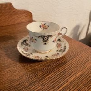 May include: A white teacup and saucer with a floral pattern and gold trim. The cup has a scalloped edge and a handle. The saucer has a matching floral pattern and gold trim.