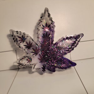 Puede incluir: Un plato de resina de color morado y transparente con forma de hoja, con purpurina incrustada en la resina. El plato tiene forma de hoja de marihuana.