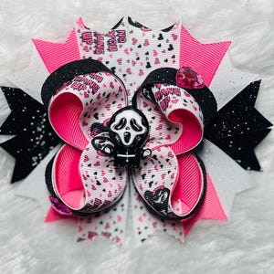 May include: A handmade hair bow featuring a black and white Ghostface character. The bow has layers of pink, white, and black ribbons, with the words "HANG UP" printed on the ribbon. A pink gem is attached to the bow.