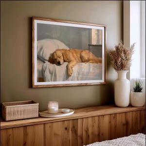 May include: A framed print depicts a sleeping golden retriever on a bed. The dog is a light golden color, resting on a white sheet and pillow. The frame is a light wood color, and the print hangs on a green wall.