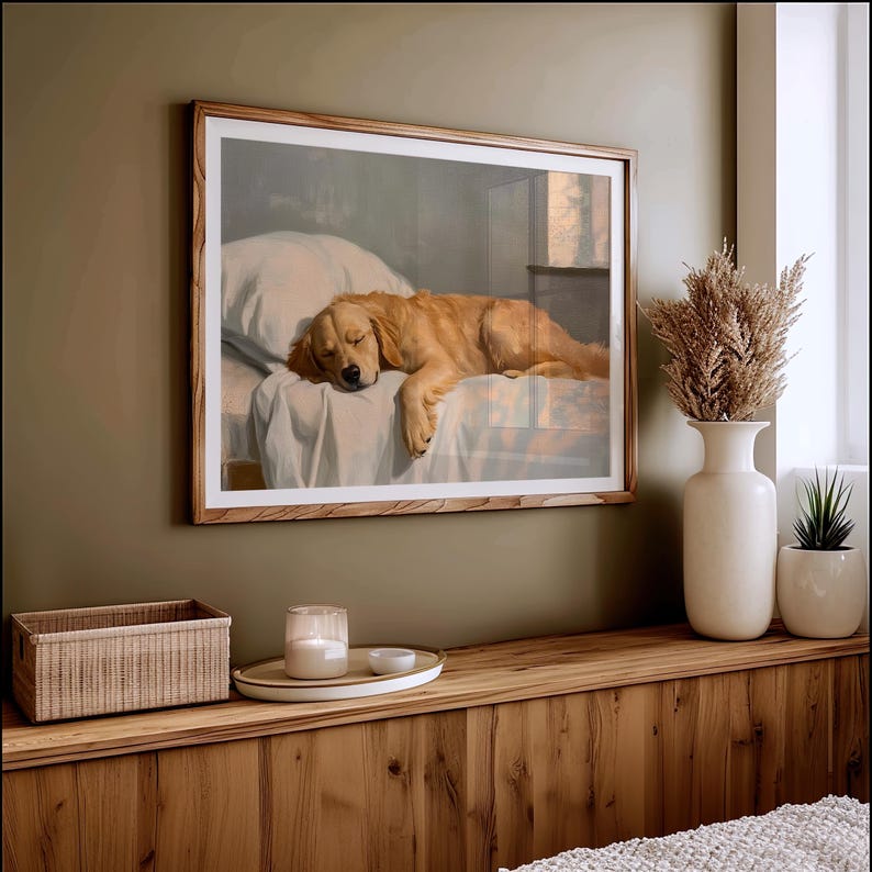 May include: A framed print depicts a sleeping golden retriever on a bed. The dog is a light golden color, resting on a white sheet and pillow. The frame is a light wood color, and the print hangs on a green wall.