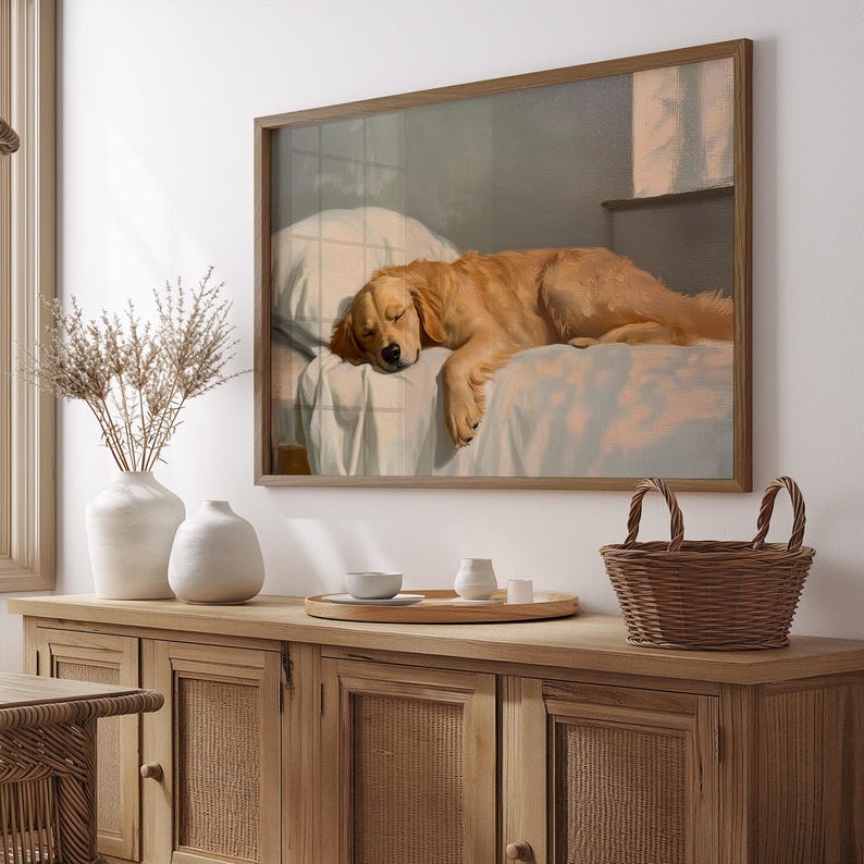 May include: A framed print depicts a sleeping golden retriever dog on a bed. The dog is light brown with closed eyes, resting on a white pillow and blanket. The frame is a light brown color. The print is displayed above a wooden cabinet.