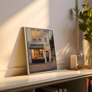 May include: A framed painting depicting a bakery with a black awning and illuminated windows. The artwork is in a silver frame, leaning against a white wall. A lit candle and greenery are in the background.