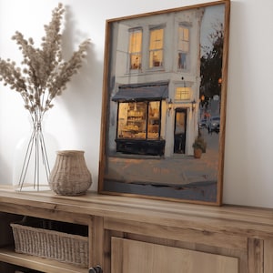 May include: A framed art print of a bakery shop at dusk. The painting shows a two-storey building with lit windows and a dark awning over the shop's display. The artwork is in a wooden frame and rests on a wooden cabinet.