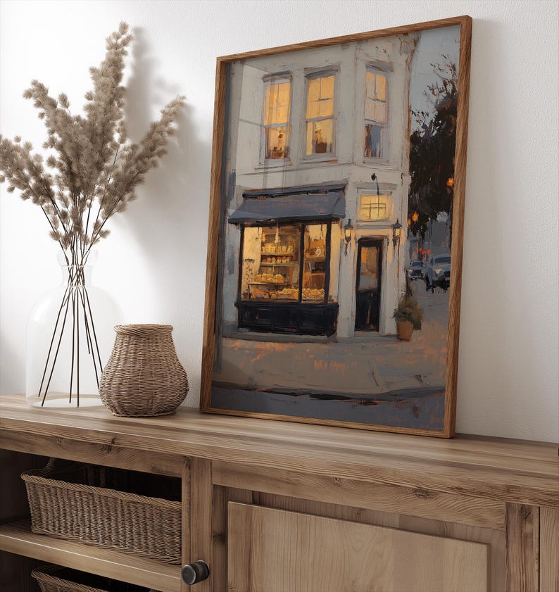 May include: A framed art print of a bakery shop at dusk. The painting shows a two-storey building with lit windows and a dark awning over the shop's display. The artwork is in a wooden frame and rests on a wooden cabinet.