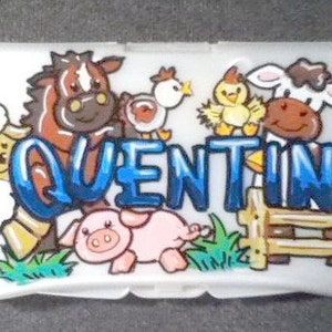 May include: A white plastic box with a farm animal theme. The box has a blue and white name "QUENTIN" written on it. The animals include a brown horse, a white sheep, a pink pig, a white cow, and two yellow chicks.