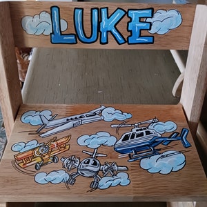 May include: A wooden children's chair painted with a blue sky and clouds. The chair is personalized with the name "LUKE" in blue letters. The seat of the chair is decorated with a yellow airplane, a blue helicopter, and a gray airplane.