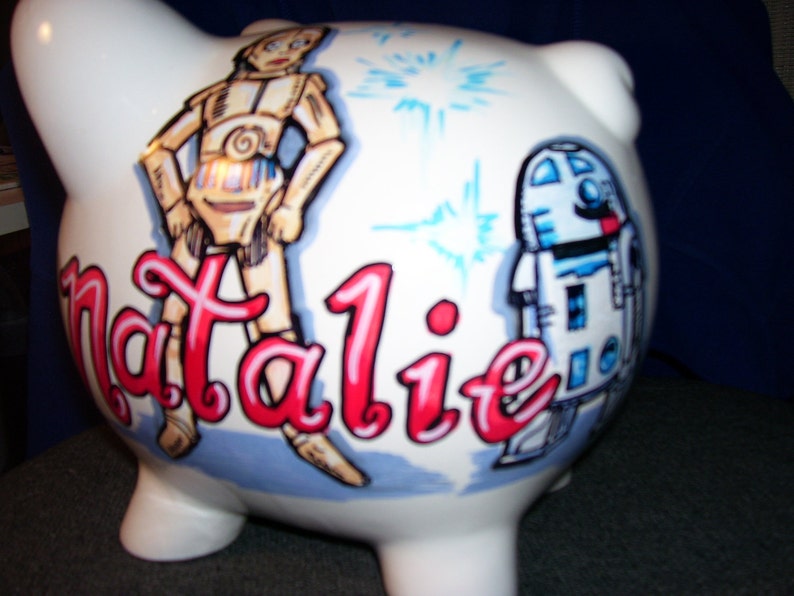May include: White ceramic piggy bank painted with a Star Wars theme. The bank features a cartoon image of C-3PO and R2-D2, along with the name "Natalie" in red lettering.