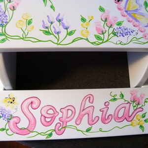 May include: White wooden step stool with a hand-painted design of pink, yellow, and purple flowers, green vines, and a butterfly. The name "Sophia" is painted in pink cursive on the step.
