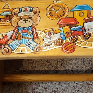 May include: A wooden step stool with a painted design of a teddy bear conductor, a train, and a clock. The stool has a built-in toilet paper holder.