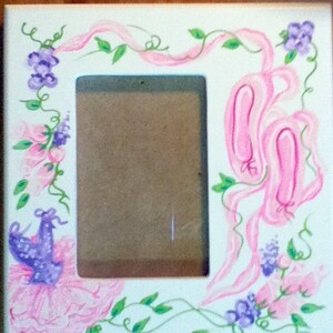 May include: White square picture frame with hand-painted ballet-themed artwork. The frame features pink ballet shoes, ribbons, and floral accents in pink, purple, and green. A purple tutu is also painted on the frame.