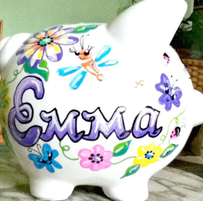 Peut inclure: Tirelire en c&eacute;ramique blanche peinte avec des fleurs violettes, des feuilles vertes, un papillon bleu et le nom "Emma" en violet.