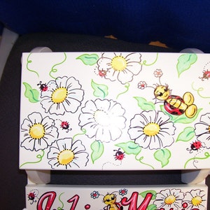 May include: A white wooden step stool with a hand-painted design of daisies, ladybugs, and a bee. The design is in black, red, yellow, and green.