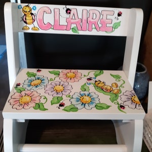 May include: White wooden step stool with a pink painted name "CLAIRE" on the back. The front of the stool is painted with a floral design featuring yellow, pink, blue, and green flowers, leaves, and ladybugs.