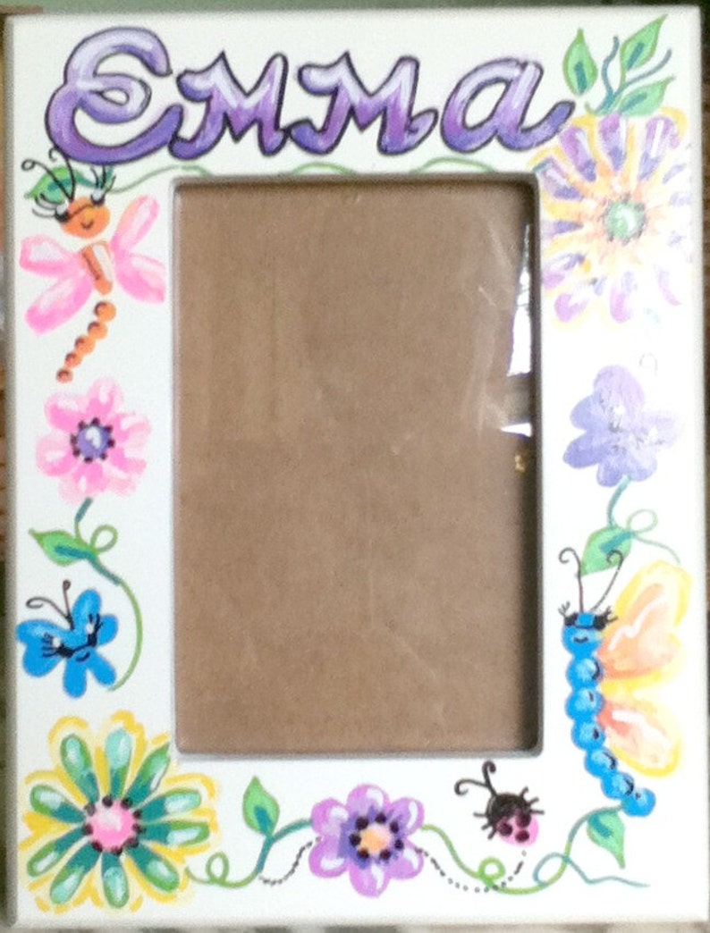 Peut inclure: Cadre photo blanc avec le nom "Emma" peint en violet en haut. Le cadre est d&eacute;cor&eacute; de fleurs, de papillons et de coccinelles peints dans diff&eacute;rentes couleurs.
