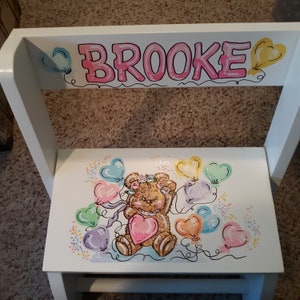 May include: White wooden step stool with a hand-painted design. The stool features the name "BROOKE" in pink letters, along with a teddy bear holding a heart and colorful heart-shaped balloons. The design includes pastel colors.