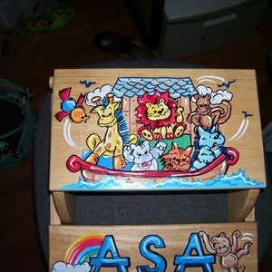 May include: A wooden children's stool with a colorful painted scene of animals on a boat. The animals include a giraffe, a lion, a monkey, a hippopotamus, and a cat. The stool is personalized with the name "ASA" in blue letters.