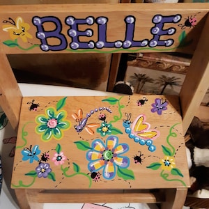 May include: A wooden children's chair painted with the name "BELLE" in purple letters with white polka dots. The seat is decorated with a whimsical floral design featuring butterflies, dragonflies, and ladybugs.