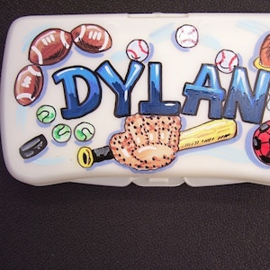 May include: A white plastic container with a blue, white, and red design. The container has the name "DYLAN" written in blue letters. The container is decorated with images of baseballs, footballs, a basketball, a baseball glove, a bat, and a soccer ball.