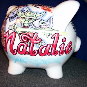 May include: White ceramic piggy bank with a hand-painted design featuring a green alien character, a blue and white character, and the name "Natalie" in red cursive.