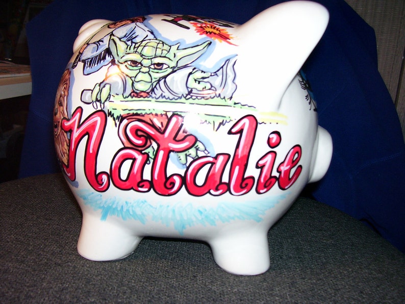May include: White ceramic piggy bank with a hand-painted design featuring a green alien character, a blue and white character, and the name "Natalie" in red cursive.