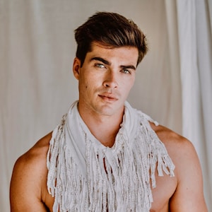 May include: A man with short brown hair is wearing a white fringed scarf around his neck. He is shirtless and has his hands clasped in front of him.