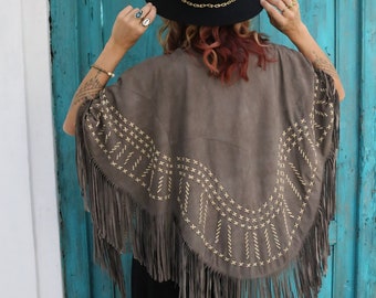 May include: A brown suede poncho with fringe and embroidered details. The poncho is worn over a black dress and the person is wearing a black hat.