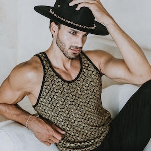 May include: A black hat with a gold chain detail. The person is wearing a black and gold geometric patterned tank top and black trousers. The person is sitting on a white surface.