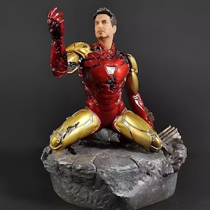 May include: A red and gold Iron Man action figure in a kneeling pose with a damaged suit. The figure is on a grey rock base.