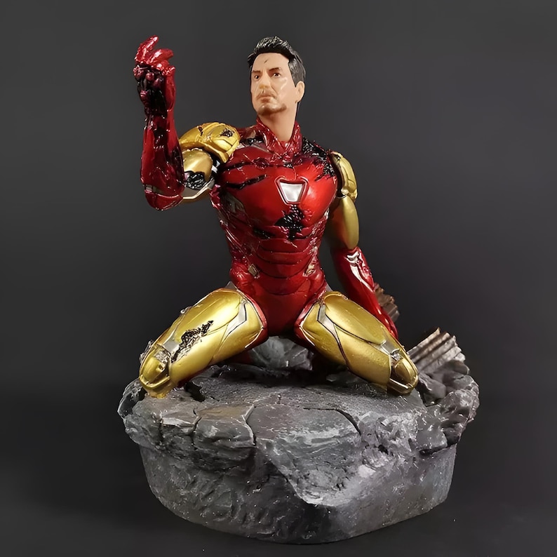 May include: A red and gold Iron Man action figure in a kneeling pose with a damaged suit. The figure is on a gray rock base.