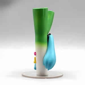 May include: A figurine of a leek with a light blue ponytail and small pink and yellow accents. The leek is primarily white and green, with a gradient effect. The figurine is standing on a clear, circular base.