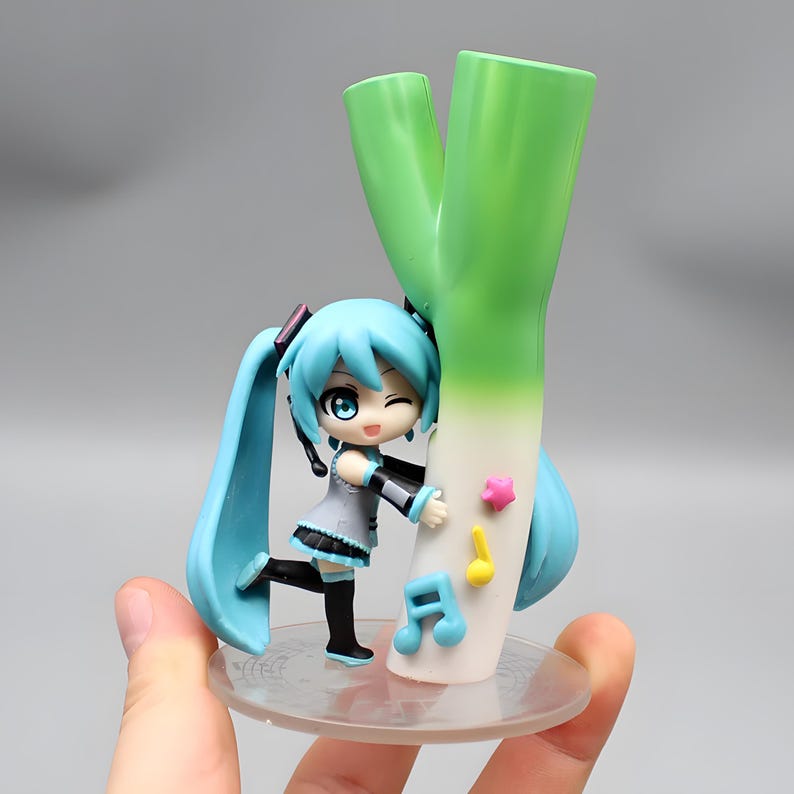 May include: A figurine of a blue-haired anime character winking and hugging a large green leek. The figure is wearing a grey dress with black accents and black boots. Decorative musical notes and a star are attached to the leek.