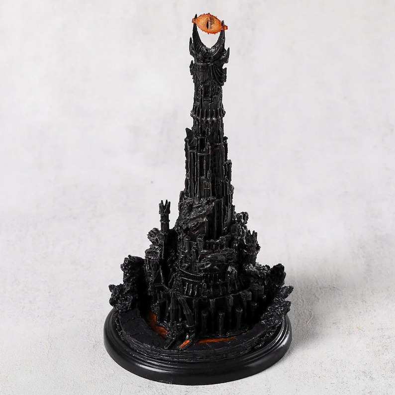 May include: A detailed black sculpture of a tall tower with a fiery orange eye at the top. The tower has intricate architectural details and sits on a round black base. The sculpture is a collectible item.