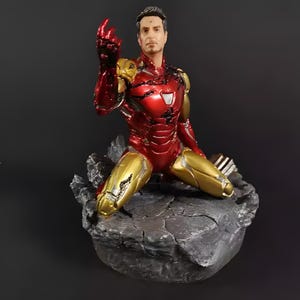 May include: A red and gold Iron Man action figure in a kneeling pose with a damaged left hand. The figure is on a gray rocky base.