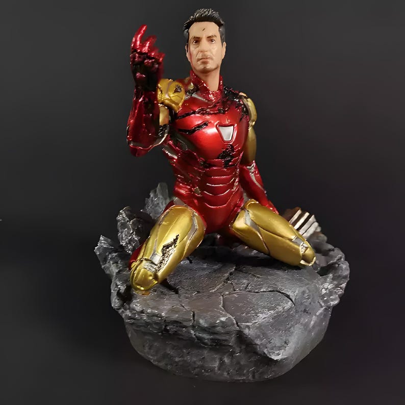 May include: A red and gold Iron Man action figure in a kneeling pose with a damaged left hand. The figure is on a gray rocky base.