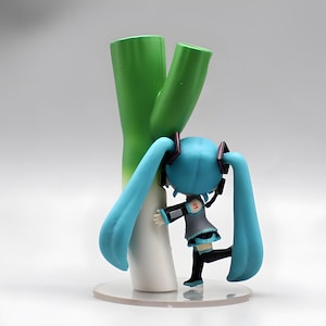 May include: A figurine of a character with long, light blue pigtails, wearing a gray and black outfit, hugging a large green and white leek. The figurine is on a clear, round base.