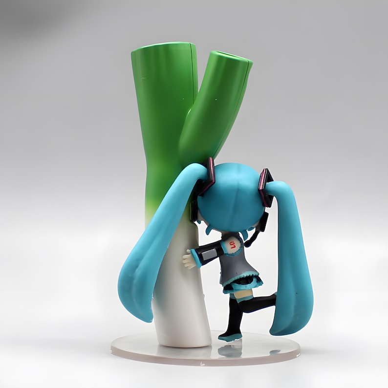 May include: A figurine of a character with long, light blue pigtails, wearing a gray and black outfit, hugging a large green and white leek. The figurine is on a clear, round base.