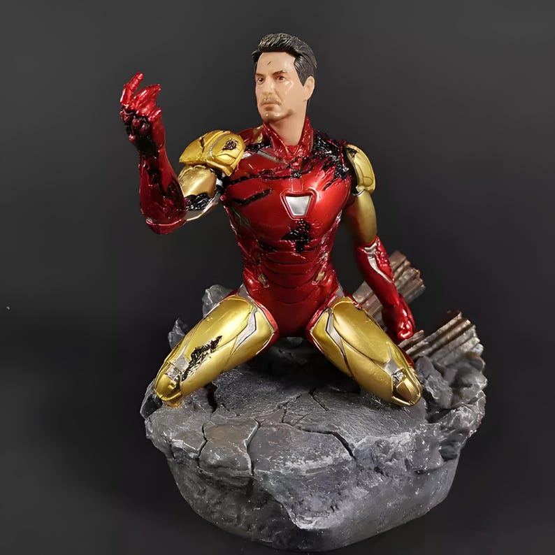 May include: A red and gold Iron Man action figure in a kneeling pose with a damaged suit. The figure is on a gray rock base.