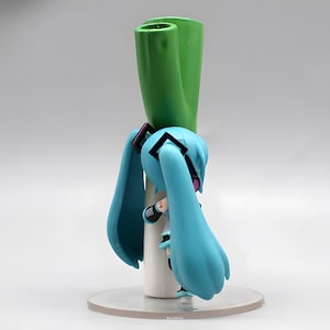 May include: A figurine of a character with long, light blue pigtails, wearing a white and black outfit. The figure is holding a large green leek. The figure is on a clear, circular base.