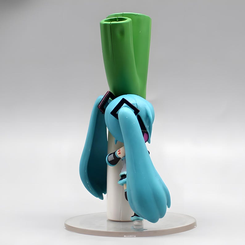 May include: A figurine of a character with long, light blue pigtails, wearing a white and black outfit. The figure is holding a large green leek. The figure is on a clear, circular base.